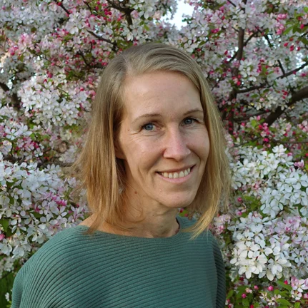 die Therapeutin Kati schaut freundlich und steht im Garten unter einem Kirschbaum der weiße Blüten trägt