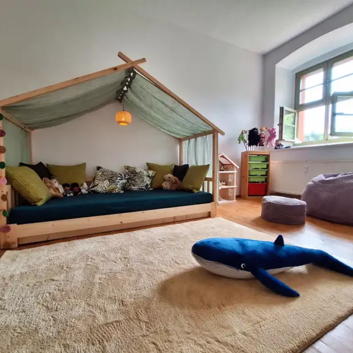 Im Kindertherapieraum ist ein Regal mit Spielzeug, ein Sessel, Sitzkissen, ein Himmelbett aus Holz und viele hübsche Stofftiere zu sehen. Wenn man aus dem Fenster schaut sieht man grüne Natur