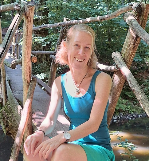 Die Traumatherapeutin Kati sitzt in der Natur auf einer Holzbrücke an einem Bach und schaut freundlich - Über Mich