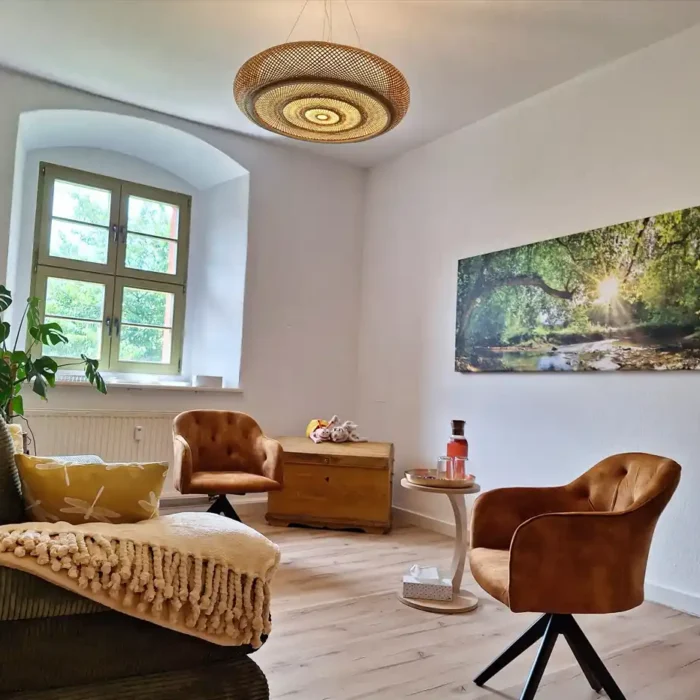 Der Therapieraum ist schön hell. Es sind eine grüne Stoffcouch, 2 bequeme bräunliche Sessel, ein Tisch mit Wassergläsern. In der Ecke steht eine Holztruhe. An der Wand hängt ein Waldbild. Durch das Fenster seht man grüne Natur. Alles ist liebevoll dekoriert und eingerichtet.