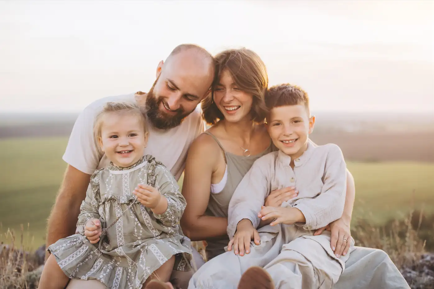 Eine Familie bestehend aus Mutter, Vater, Tochter, Sohn sitzen zusammen in der Natur auf einem Hügel. Die Kinder sitzen auf dem Schoß der Eltern. Die Stimmung ist vertraut und alle schauen zufrieden.