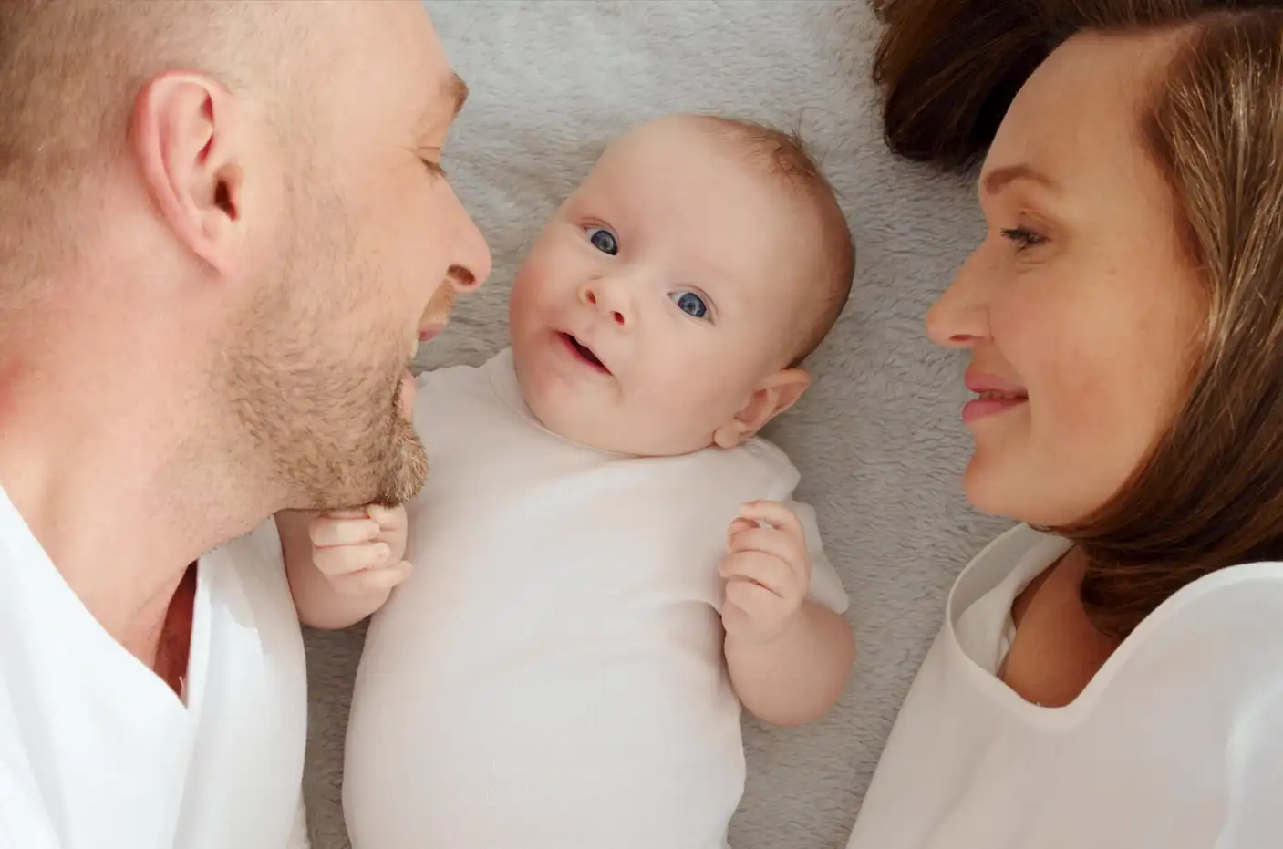 Ein Baby liegt zwischen seinen glücklichen Eltern im Bett und schaut zufrieden und neugierig. Symbolbild für Säuglinge